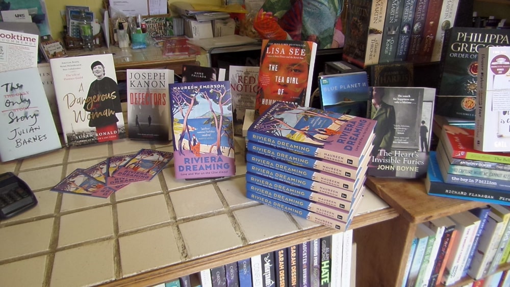 Display of paperback novels on a tiled bookstore counter, with multiple copies of Riviera Dreaming visible and promotional cards nearby.